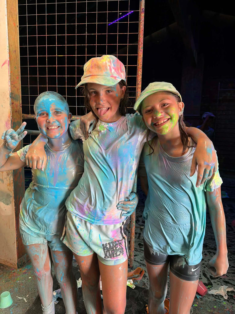 Three children with colorful paint just outside the Splatter & Splash room showing their faces and clothes, posing together.